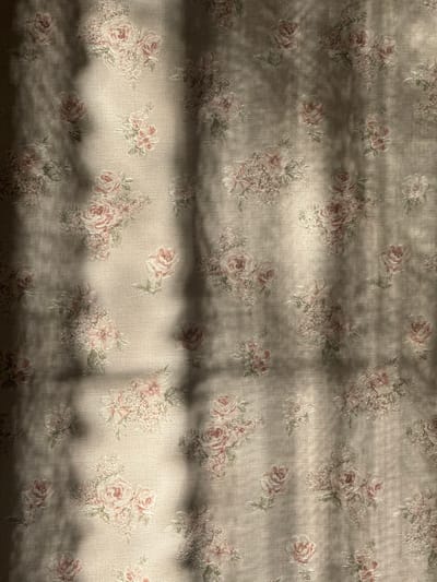 This image depicts a floral patterned fabric, likely a curtain or wallpaper, illuminated by natural light in an interior setting within the city of Zelenogradskiy, Russia. The fabric background is a light beige or cream color, patterned with clusters of pink and rose-colored roses, light green leaves, and smaller, intricate floral elements. The entire surface is crisscrossed by distinct, irregular shadows, indicative of sunlight filtering through an external obstruction such as window mullions or foliage. No individuals, direct actions, or other significant objects are visible within the frame. The interaction involves direct sunlight casting sharp shadows onto the patterned textile surface.