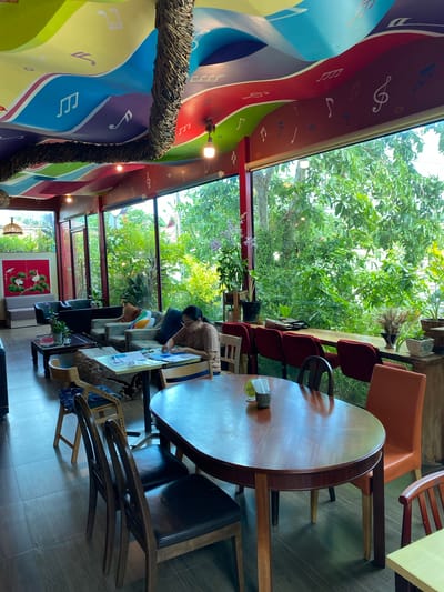 This is an interior photograph of a cafe in Tambon Bang Sare, Thailand. The cafe features a vibrant, wave-patterned ceiling adorned with musical notes and musical symbols. Large windows offer a view of lush greenery, suggesting a garden or outdoor seating area.
In the foreground, a large oval wooden dining table is set with several chairs. Two chairs are dark brown with black seats. An orange chair is visible to the right. On the table, there's a small metal container with napkins and a small coaster with writing on it.
In the middle ground, a woman with glasses is seated at a lighter colored table, appearing to be working or reading. She is wearing a light brown t-shirt. In front of her are papers and a pen. Several plush armchairs and sofas are arranged in the background, along with coffee tables. On one coffee table, there are papers, a cup, and some decorative items. A large red artwork featuring birds and branches hangs on the wall.
The flooring is a dark tiled surface. The overall ambiance suggests a relaxed and artsy environment.