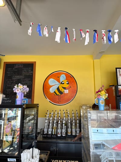 This media file captures a bright and cheerful interior scene, likely a cafe or coffee shop. The dominant color is a vibrant yellow on the walls, which creates a warm and inviting atmosphere.
In the foreground, a string of small, colorful flags made of fabric hangs diagonally from the left. These flags appear to be red, white, and blue, giving a festive or patriotic feel. Further to the left, a metallic track or rail system is visible, suggesting a functional element of the space, possibly for doors or equipment.
The center of the image features a whimsical painted mural of a bee on a yellow wall, with the bee holding a small cup. This artwork adds to the playful character of the establishment. Below the mural, a dark chalkboard displays a menu of drinks, with sections for "LATTES," "LEMONADES," and various drink names like "HONEY BEE," "QUEEN BEE," "PINK MANGO," and "PURPLE BREEZE." Some of the drink names are creatively related to bees, reinforcing the theme from the mural. A small bouquet of artificial purple, white, and yellow flowers is placed on top of the chalkboard.
To the right, a counter area is visible with a refrigerated display case filled with various items, including what looks like pastries or desserts. Above the display case is a rack holding numerous bottles of flavored syrups, identified as "MONIN." A hand is reaching towards one of these bottles, suggesting someone is preparing a drink. In the background, there are more shelves with bottles, possibly spirits, indicated by a visible "BACARDI" label. A stack of white napkins is also present on the counter. The overall impression is a busy but friendly service environment.