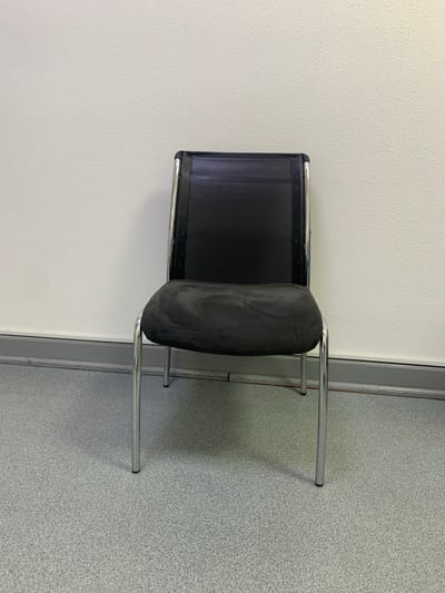 A black mesh-backed chair with a dark fabric seat and chrome legs sits on a speckled gray floor in front of a textured white wall. A gray baseboard runs along the bottom of the wall. The scene is indoors and appears to be a neutral, possibly institutional or waiting area environment. There are no people or actions depicted in the image. The lighting is even and appears to be from overhead. The location is not specified within the image itself.