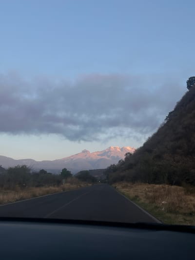 The image shows a mountain range, possibly near Santiago Xalitzintla, Mexico, partially obscured by clouds. The sky is light blue transitioning into the grey of the clouds. A road runs along the right side of the frame. Vegetation is visible along the roadside. The photo appears to have been taken from inside a vehicle, as a portion of the vehicle's interior is visible on the right. The mountain peak is highlighted by sunlight.