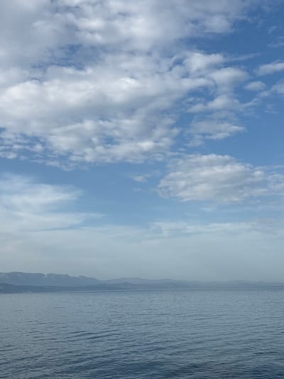 The image shows a body of water, possibly a lake or sea, under a sky with scattered clouds. The water is dark gray and appears rippled. In the distance, a hazy shoreline with mountains is visible. The sky transitions from clear blue with white clouds on the left to a misty gray haze on the right. The image does not contain any people. The location is unknown, and there is no evidence the image was taken in Bondarenkovo, Ukraine.