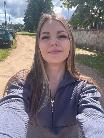 A person, likely female, is taking a selfie outdoors. She has long, light brown hair with blonde highlights. She is wearing a textured, blue-gray zippered jacket. The background shows a dirt road and a green vehicle, possibly a jeep. A wooden fence and some buildings can be seen in the distance, suggesting a rural or semi-rural setting. Judging from metadata, the image was taken in Slobodka, Russia.