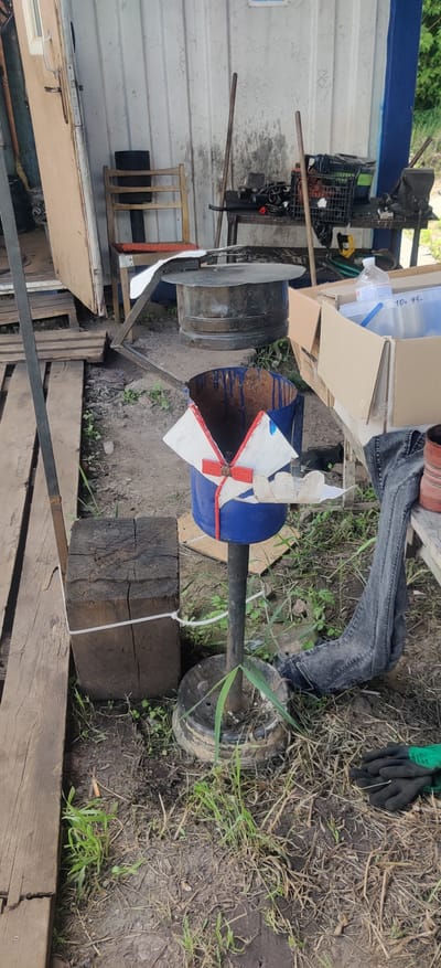 The image depicts an outdoor scene in Rizunenkove, Ukraine, likely during daytime under overcast or moderate lighting conditions. The primary subject is a homemade device resembling a wind turbine or a fan, constructed from a blue barrel with white and red blades attached to a central rotating shaft. This shaft is mounted on a stand that appears to incorporate a wheel, suggesting it might be mobile or intended for outdoor use.
Beside the wind turbine, a large, dark metal pot or cauldron is visible, positioned on a metal frame. In the background, there are various objects including cardboard boxes, a plastic bottle, a black plastic crate, and what seems to be a pair of green work gloves lying on the ground. The ground is covered with dirt and dry grass. The scene also includes wooden elements, such as planks and a thick log, which seem to be part of the surrounding environment or supporting structures.
A person's legs, clad in dark denim jeans and possibly wearing work boots, are visible on the right side of the image, suggesting human presence and activity. The overall setting appears to be a workshop, yard, or a rural area. There is no clear indication of a specific event taking place, but the arrangement of objects suggests ongoing work or a completed project. The image is oriented sideways, with the top of the frame pointing to the left. No visible text is discernable.