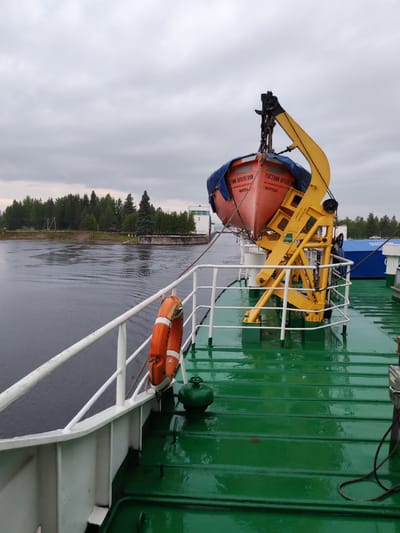 The image shows the deck of a vessel, with a lifeboat suspended by a yellow davit. The lifeboat is orange with a blue tarp covering its top. Text on the lifeboat is in Russian, reading "ЕВДЕНИИ ОРЛОВСКИМ" and "КОТЛАС". There is a white railing along the edge of the deck, and a life preserver hangs from it. The deck itself is painted green and appears wet, reflecting the cloudy sky. In the background, there is a body of water, a line of trees, and a building with a "2" on it. The overcast sky suggests a cool, possibly damp, day. The image does not provide specific information about the city of Belousovo, Russia.