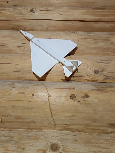 A white wooden model of a supersonic jet, resembling the Concorde, is mounted on a wall made of horizontal wooden logs. The model is positioned in the upper left quadrant of the image, with its nose pointing towards the top left. The jet has a delta wing shape and twin vertical stabilizers at the rear. The logs of the wall exhibit a natural wood grain and color variations, with visible knots. The lighting suggests an indoor environment. The specific location in Молочное, Russia, is not determinable from the image alone.