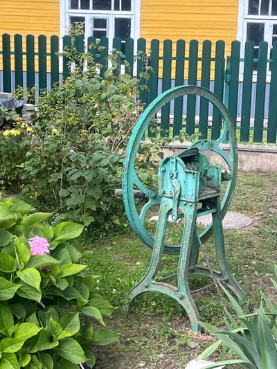In Zhaludok, Belarus, a turquoise metal contraption, likely a fodder cutter, stands in a grassy yard. The machine has large spoked wheels and a complex mechanism of gears and blades, showing signs of rust and age. It is positioned near a vibrant green wooden fence, which separates the yard from a building with a bright yellow wooden exterior and white-trimmed windows. Lush green foliage, including large-leafed hydrangeas with a single pink bloom, surrounds the machine and fence, suggesting a garden setting. The ground is covered in a mix of grass and dirt.