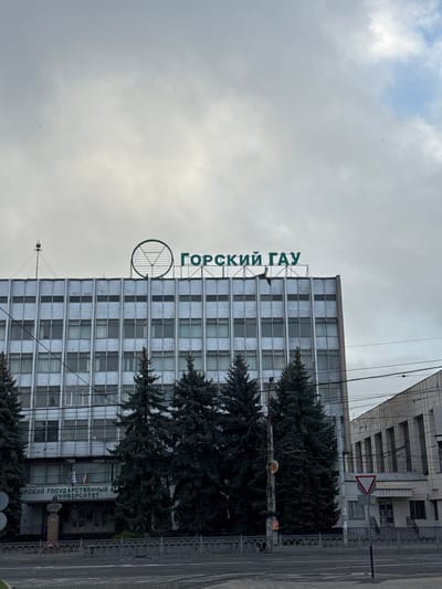 The image shows a building in Akhieli, Georgia. The building has a sign on the top that reads "ГОРСКИЙ ГАУ" in green letters. Below that, there is a round symbol. There are trees in front of the building. A road and a sidewalk are on the right side of the image. The sky is overcast. A sign with a red border stands at the street corner.