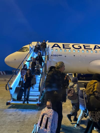 People are disembarking an Aegean A320neo aircraft via stairs. Several passengers, including some with luggage, are visible on the steps. The plane's fuselage, with "AEGEAN" printed in blue, is partially visible in the background, along with a blue sky. The image is taken from a close perspective, capturing the moment of arrival or departure.