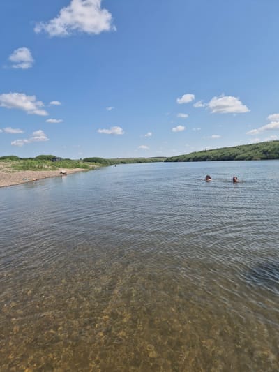 The image depicts a wide, calm river under a bright blue sky dotted with white clouds. The river's bottom is visible in the foreground, revealing a bed of rocks and pebbles. On the left bank, there's a gravelly shore with sparse green vegetation and a dark-colored vehicle parked near the edge. Further back, the landscape is covered in dense green bushes and trees, forming a natural embankment that curves with the river. In the middle of the river, two individuals are swimming, with their heads and shoulders visible above the water. One swimmer appears to be wearing an orange flotation device. The overall scene suggests a peaceful, outdoor recreational setting in the rural area of Novo-Ishim Rural District, Kazakhstan.