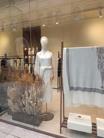 The image shows a storefront window display in Molokovo, Russia, featuring a mannequin dressed in white knit clothing, including a dress and a shawl. The mannequin is accessorized with a white headband and is positioned in front of a display of dried reeds. To the right, a large, light-colored textile with a grey border and intricate pattern, possibly a blanket or shawl, is hung on a wooden rack. Below the textile, on a shelf, is a white gift box with the brand name "ORENSHAL" printed in grey, along with Russian text indicating "FABRIKA ORENSBURGSKIH" and "PUSHOVYKH PLATKOV" (Orenburg Down Scarves Factory). In the background, a rack of clothing is visible, and the reflection of an ornate building interior, likely the GUM department store in Moscow, suggests the location might be within a larger shopping complex rather than a standalone store in Molokovo itself. The lighting is neutral, and the overall scene conveys a sense of retail display and product presentation.