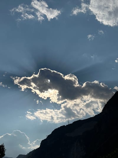 This is a vertically oriented, close-up photograph of a dramatic sky with sun rays shining through clouds. The main subject is a dark, dense cloud positioned centrally, with its edges brightly illuminated by the sun. Sunbeams emanate from behind this cloud, creating a "god ray" effect. To the right of the frame, a dark, imposing cliff face or mountainside occupies the majority of the space. The sky is a clear blue with scattered, wispy white clouds visible in the upper left and lower right portions of the image. The lighting suggests it is daytime, likely late afternoon or early evening due to the angled sun rays and the shadows cast by the cliff. The overall mood is one of awe and natural beauty, highlighting the dynamic interaction of light and cloud. There are no people or text visible in the image. The location context of Cividate Camuno, Italy, suggests this may be a mountainous or hilly region.