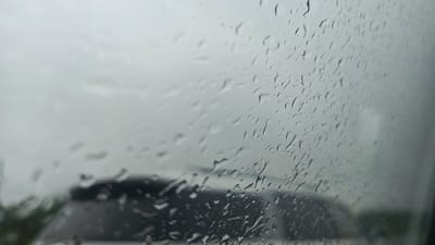 A close-up, low-angle, exterior shot captures a car's windshield during a rain shower. The windshield is covered in numerous water droplets of varying sizes and shapes, creating a distorted view of the outside environment. The background is blurred due to the rain and the focus on the droplets, but hints of a dark-colored vehicle and some greenery are discernible. The overall lighting suggests an overcast day. The image is taken from within a vehicle in Lesnye Morkvashi, Russia.