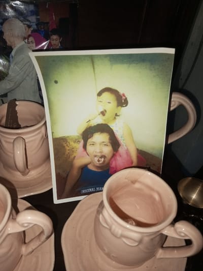 A photograph of a smiling child and adult with treats on their faces stands among a set of pale pink tea cups and saucers. The cups are mostly empty. A small brass spoon sits beside one cup. Other photographs are visible in the background, partially obscured by the tea set. The scene is dimly lit, creating a slightly nostalgic atmosphere.