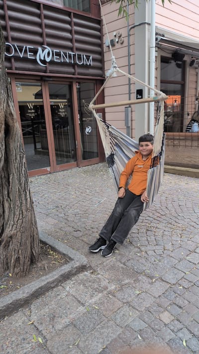 A young boy is relaxing in a hammock suspended from wooden poles. He is wearing an orange hoodie with the word "World" printed on it, dark-colored pants, and black sneakers. The hammock has a striped pattern of white, blue, and black. The boy is lying back with his head resting on the hammock and his eyes open, looking towards the camera.
The scene is outdoors, with a cobblestone ground covered in fallen leaves, suggesting it might be autumn. Behind the boy is a building with large glass doors. One of the doors has signage visible, including "BAR & FITNESS" and a logo. Another sign on the building reads "OVE MENTUM". The building's facade is a combination of corrugated metal and glass. To the left of the building, there are some plants. The lighting suggests it is daytime, possibly late afternoon due to the angle of the shadows. The boy appears relaxed and content. The location is Östersund, Sweden.