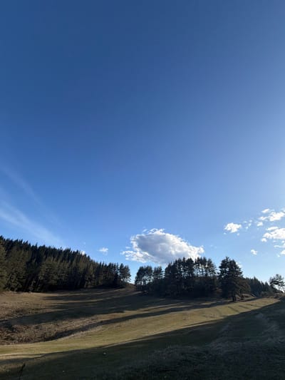 The image depicts a rural, forested landscape under a clear blue sky, located in Kasak, Bulgaria. The upper two-thirds of the frame are dominated by a deep blue sky with sparse white cirrus clouds in the upper left and a prominent, brighter cumulus cloud near the center-right. The sun, positioned to the right, casts a bright light on the right side of the sky. The lower third of the image features a gently undulating grassy hillside, largely dry with patches of greener vegetation. Long shadows extend from left to right across the hillside, indicating a low sun angle. A dense line of dark green coniferous trees, likely pine or fir, forms the horizon, covering slopes that rise towards the left and right. No people or animal subjects are visible. No specific actions or interactions are occurring within the frame. The environment is characterized by natural terrain, dense forestation, and open grassland.