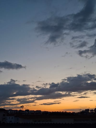 The image shows an orange sunset viewed from Paralimni, Cyprus. A line of buildings is silhouetted against the sunset's orange glow with a small windmill visible in the city skyline. The sky is filled with clouds of varying shades of grey. The darker clouds give way to the blue sky behind them.