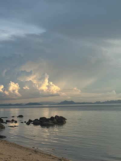 The image depicts a serene coastal scene. The foreground features a sandy beach transitioning into the calm, reflective waters of the sea. Several dark, wet rocks are visible along the shoreline and partially submerged in the water, some forming a small cluster. In the distance, a hazy silhouette of mountains lines the horizon. The sky is dramatic, with large, fluffy cumulus clouds tinged with yellow and white from sunlight breaking through, juxtaposed against a backdrop of overcast, grey clouds. Rays of sunlight are visible piercing through the clouds, creating a celestial effect. The overall impression is one of natural beauty and tranquility. This scene is located in Baan Pang Ka, Thailand.