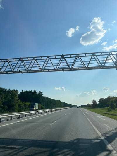 This media file captures a bright, sunny day on a highway in Verkh-Tula, Russia. The scene is dominated by a clear blue sky with scattered white clouds, suggesting fair weather. On the left side of the image, a large, metallic overhead structure, likely a gantry for road signs or traffic lights, spans across the frame. Its geometric, truss-like design is prominent.

To the right of the gantry, a multi-lane asphalt highway stretches into the distance, bordered by lush green trees and vegetation on either side. The road appears to be well-maintained with white lane markings visible. A few vehicles are seen on the highway, including what appears to be a truck in the distance. Shadows from the overhead structure and possibly the vehicle from which the photo was taken are cast across the road, indicating the sun is high in the sky, likely mid-day.

The overall impression is one of travel and open road, with the vast sky and endless highway creating a sense of freedom. The image is oriented vertically, suggesting it was taken from a moving vehicle looking upwards and forwards. No people are clearly visible, but the presence of vehicles implies human activity. There is no visible text in the image.