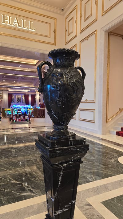 A dark marble vase with ornate carvings sits on a matching marble pedestal in the foreground. The vase has two handles and a wide mouth, decorated with what appear to be mythological figures or cherubs. The pedestal is square with decorative molding at the top. The floor is made of dark, polished marble with intricate veining. In the background, the scene opens to a brightly lit interior that resembles a casino or hotel lobby. Rows of slot machines are visible, along with people seated and walking around. The walls in the background are cream-colored with gold trim and decorative paneling. A sign above an entrance reads "HALL" in illuminated gold lettering. A staircase with a red carpet runner and gold railing is visible on the right side of the background. The overall impression is one of opulence and grandeur. The location of Agios Epiktitos, Northern Cyprus / Cyprus is not discernible from the image.