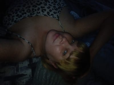 An individual is lying on a bed with a leopard-print top. The individual's head is turned to the side, and their eyes are looking at the camera. The background is dark and indistinct, suggesting an indoor setting, possibly a bedroom. The image is taken from a high angle, looking down at the individual. The individual appears to be wearing makeup, and their hair is blonde. The lighting is dim, with a spotlight illuminating the individual's face. The overall impression is one of intimacy and casualness. There is no specific information in the image to indicate the location in the city of Sibircevo, Russia.