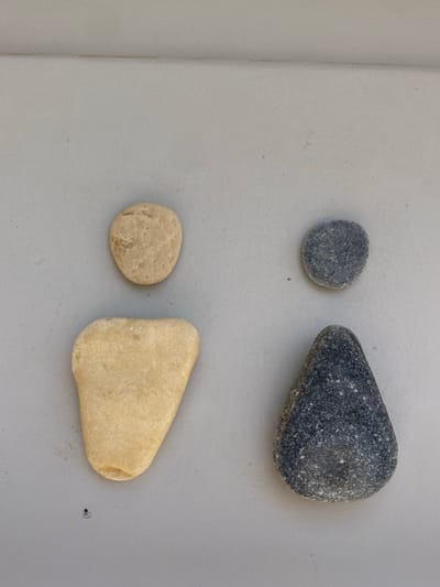 A static scene displays four natural stones arranged into two distinct anthropomorphic figures on a light gray surface, reportedly located in Φάρος, Greece. On the left, a light beige, oval stone (head) is positioned above a larger, rounded, irregular light beige stone (body). On the right, a dark gray, speckled oval stone (head) sits above a larger, teardrop-shaped dark gray speckled stone (body). All stones are smooth and weathered. The background is a uniform, flat light gray surface, possibly a wall or floor, showing a subtle horizontal line in the upper area. Soft shadows beneath the stones indicate they are resting on the surface. No human individuals or active interactions are depicted.