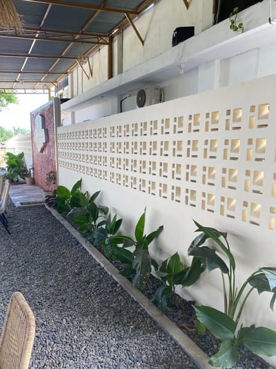 The image depicts an outdoor seating area, likely part of a cafe or restaurant, in Aluepiet, Indonesia. A white decorative wall with geometric cutouts runs along the side of the area, separating it from a gravel ground cover. Lush green plants are planted along the base of the wall. In the background, a corrugated metal roof is supported by a metal frame, suggesting an open-air structure. A sign with text, partially obscured by a wall, is visible on the left. The overall impression is one of a relaxed, tropical ambiance.