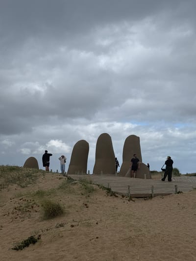 The image displays the iconic "La Mano" (The Hand) sculpture in Punta del Este, Uruguay, on a cloudy day. Five large, finger-like structures made of concrete or similar material emerge from a sandy beach, stretching towards the left side of the frame. The sculpture appears to be set within a sandy dune environment, with sparse green vegetation visible on the right side.
Several people are present around the sculpture. On a wooden boardwalk running parallel to the fingers on the right, one person in dark clothing and a hat stands near the outermost finger, holding what might be a stick or tool, looking towards the left. Closer to the viewer, another individual in a dark shirt and light shorts stands next to the second finger, looking forward. Further down the line of fingers, two more people are visible: one in a light top and dark pants near the third finger, and another in dark attire near the fourth. One more person is partially obscured behind the fifth finger. They appear to be tourists, observing the sculpture or posing for photos.
The sky occupies the upper portion of the image and is heavily overcast with shades of grey, indicating a cloudy or potentially rainy day, though there are no signs of precipitation. Despite the clouds, it is clearly daytime. The sandy beach stretches into the foreground on the right, with a wooden pathway guiding visitors along the sculpture. There is no visible text in the image.