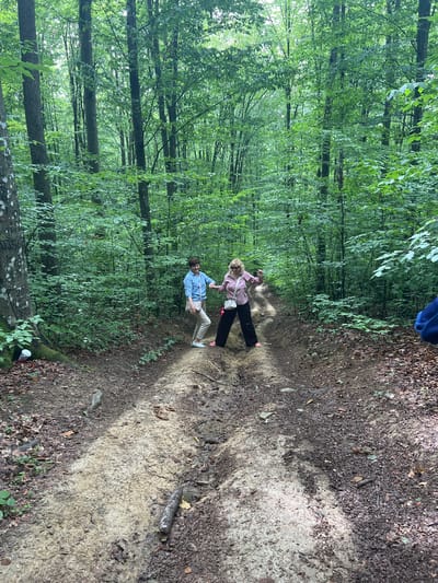 Two individuals, a male and a female, are captured standing on a dirt path in a dense forest. The male, on the left, wears a blue shirt and light-colored pants. The female, on the right, is dressed in a pink shirt and dark pants, with sunglasses perched on her head. They appear to be interacting with each other or posing for the photo. The path is uneven, with signs of mud and debris, suggesting it may be a hiking trail or a less-maintained road. The surrounding environment is characterized by numerous tall trees with lush green foliage, creating a shaded and natural setting. The image was taken outdoors, likely during the daytime, given the natural light filtering through the trees. The location is identified as the city of Pavlovo, Ukraine, implying the forest is situated within or near this area.