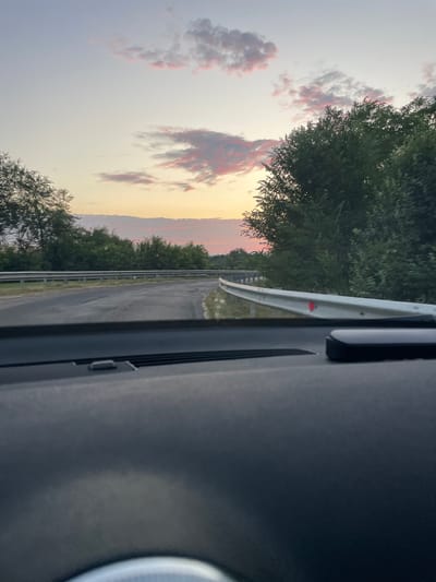 The image captures a view from inside a vehicle, looking out at a road during a sunset. The sky is painted with soft hues of pink and orange, with scattered clouds reflecting the fading sunlight. A road curves to the right, bordered by a metal guardrail on both sides. Dense green trees line the roadside, creating a natural backdrop. The dashboard of the car is visible in the foreground, partially obscuring the lower part of the scene. The overall impression is one of a tranquil drive through the Moldovan countryside near Cricova.
