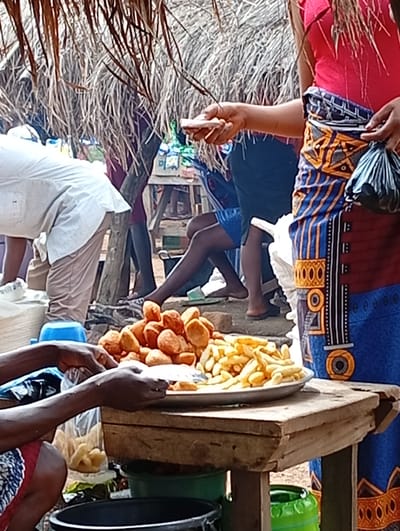 In Tse Igbudu, Nigeria, an outdoor market stall displays a variety of fried foods. The stall has a thatched roof and wooden supports. In the foreground, a person's hands are shown reaching for a plate of golden-brown fried snacks, possibly puff puff or similar dough balls, and a serving of french fries. Another person, wearing a vibrant blue and orange patterned fabric and a pink top, stands behind the stall, holding a black plastic bag. Other individuals, partially visible, are dressed in casual clothing, with one person wearing a white shirt and beige pants. The ground appears to be dirt. In the background, more market activity and structures can be seen.