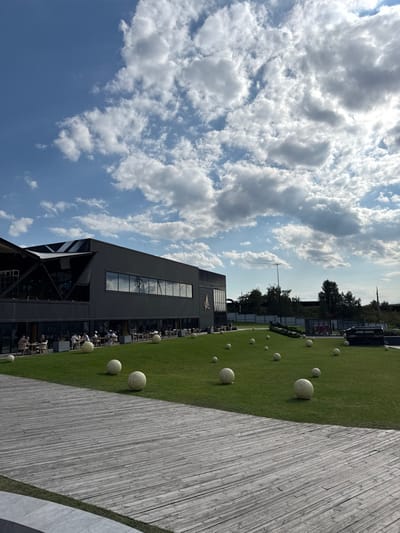 The scene depicts an outdoor leisure area at the Osocor Residence in Oseshchyna, Ukraine, under a partly cloudy sky. A multi-story, dark-colored building with large reflective windows and an integrated patio area occupies the mid-ground left. Approximately twenty individuals are seated at tables on this patio, engaged in static activities such as dining or conversation. A stylized avian emblem is affixed to the building's right facade. A manicured green lawn spans the area between the building and the foreground, populated by approximately twenty spherical, light-colored decorative objects, varying in diameter from 0.3 to 0.8 meters. The immediate foreground features a wide boardwalk constructed of horizontally laid, weathered wooden planks. In the distant background, beyond a treeline, a tall utility pole and indistinct industrial structures are visible.