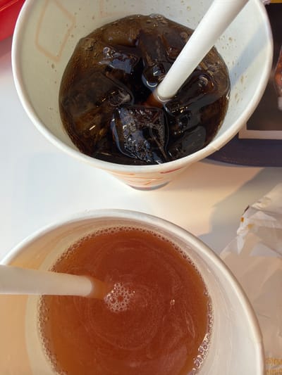 A top-down, close-up view shows two beverages in white paper cups on a light-colored surface, characteristic of a fast-food or casual dining setting in Chuchuligovo, Bulgaria.
The cup on the left contains a dark, carbonated drink, likely a cola, with numerous ice cubes and visible bubbles. A white paper straw is submerged diagonally into the beverage. The outer side of this cup has faint, partially visible red and orange patterned markings.
The cup on the right holds an amber-colored liquid, resembling iced tea or juice, also with a white paper straw. Small bubbles encircle the liquid's edge and gather around the straw.
In the background, a dark blue or black object, possibly a menu or tray, is partially visible behind the left cup. Above and between the cups, crumpled white paper, likely a napkin or food wrapping, is present. Faint, unidentifiable text is barely visible on this crumpled paper. The scene is brightly lit, indicating a daytime setting or a well-illuminated indoor environment. No people are visible.