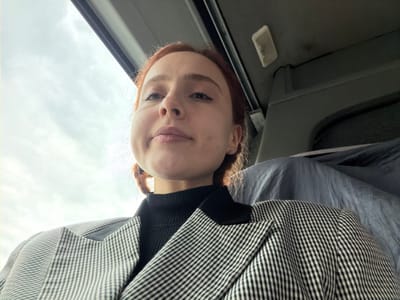 The image displays a young adult female seated inside a vehicle, captured from a low-angle perspective. She has reddish-brown hair, fair skin, and a neutral expression with slightly pursed lips, gazing upward and to the right. She is dressed in a black ribbed turtleneck beneath a black and white checkered blazer. The vehicle's interior features a light gray ceiling, with a rectangular overhead light fixture visible in the upper right corner. A dark structural element is positioned behind her head, and a light blue or gray draped fabric object is situated to her right. The upper left portion of the image reveals an overcast sky with visible clouds, seen through what appears to be a window or sunroof. The specific location within the city of Eschau, France, cannot be determined from the visual evidence provided.