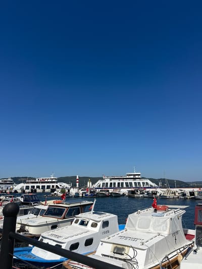 The image shows a clear blue sky above a harbor filled with various boats. In the foreground, several smaller white motorboats are docked. One boat in the center has the name "DENİZKUSÜ 17" and "17 D 1151" written on its side, along with a Turkish flag. Another boat to its left has "17 D 1570" written on it. In the background, larger ferries are moored, with one ferry displaying the logo "Gestaş." A red and white striped lighthouse is visible on the right side of the background. The boats are in the water, and the scene appears to be a busy port. The location is identified as the city of Çanakkale Merkez, Turkey.