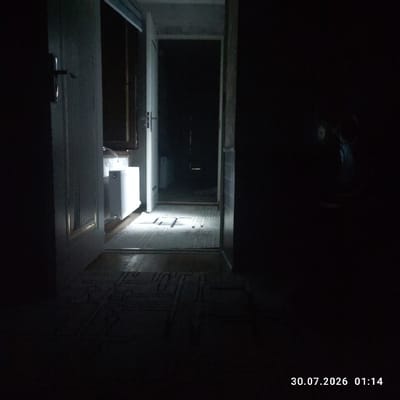 The image depicts an indoor scene with poor lighting, possibly a hallway or entryway. A white door is ajar on the left, revealing a brightly lit radiator behind it. A narrow doorway or archway is visible in the center, leading into a dark room. The floor in the foreground appears to be tiled with a patterned rug, upon which light is casting distinct shadows. In the far right of the image, a dark object, possibly a fan, is partially visible. A timestamp in the bottom right corner reads "30.07.2026 01:14". The scene does not provide any specific visual cues to pinpoint its location within Uchqoʻrgʻon, Uzbekistan, beyond it being an interior space.