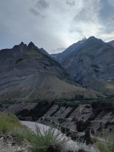 This media file depicts a scenic mountain landscape in Gushal, India, under a cloudy sky, possibly during the late afternoon or early evening. The primary subjects are the majestic, rugged mountains that dominate the frame. Their slopes are a mosaic of barren rock, sparse vegetation, and patches of green, suggesting a high-altitude, arid environment.

In the foreground, a muddy river winds its way through a valley. The riverbanks are lined with scrubby bushes and sparse green vegetation, with a dirt road or path visible snaking along the hillside adjacent to the river. The terrain is characterized by steep slopes and terraced areas, indicative of human intervention for agriculture or habitation, though no people are visible.

The sky is filled with dramatic clouds, with hints of sunlight breaking through in the upper right portion of the image, creating a soft, diffused light that casts a somber yet beautiful mood. The overall atmosphere is one of natural grandeur and serene isolation. The texture of the mountains, from the sharp peaks to the undulating slopes, is richly detailed, highlighting the geological formations. No text is visible in the media file.