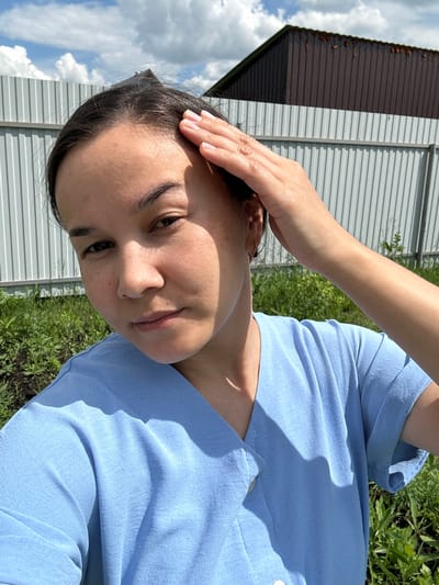 A close-up, medium shot captures a young woman outdoors on a sunny day, likely in Osorgino, Russia. She is wearing a light blue blouse and her right hand is raised, touching her forehead. Her dark hair is pulled back, and her face is turned slightly to the left, looking towards the camera. The background features a light gray corrugated metal fence, green foliage and grass, and a dark brown building with a rusted roof. The sky above is partly cloudy with white cumulus clouds against a blue expanse. The lighting suggests natural daylight, casting shadows on the woman and the surrounding environment.