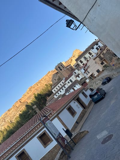 The image depicts a street scene in the town of Alpuente, Spain. The perspective is from a low angle, looking up a cobblestone street that slopes upwards. The buildings are typical of a Spanish village, with white stucco facades, terracotta tiled roofs, and some featuring stone accents.

On the left side of the street, a building with a white lower story and a stone foundation is prominent. It has at least two windows with dark, barred grills. Further up the street, on the right side, are more buildings with multiple stories, balconies, and windows. One building has a distinctive cream-colored facade with brown trim around the windows and balconies.

Dominating the background on the left is a large, steep, orange-toned rock formation, likely a cliff face or a natural geological feature. Pine trees line the base of this formation. A church tower with a clock face is visible in the distance, partially obscured by the buildings.

The street is paved with gray cobblestones. Two cars are parked or driving on the street: a dark-colored vehicle is further up, and a teal-colored hatchback is closer to the foreground. A street lamp with an ornate black metal fixture is mounted on the side of a building near the top of the image. A public information sign is situated near the foreground building on the left, and a trash receptacle is nearby. The sky is clear and blue, indicating daytime. The sun appears to be casting long shadows, suggesting it might be late afternoon or early morning.