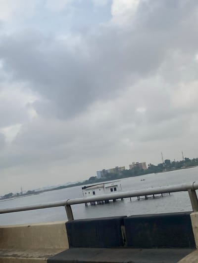 The scene depicts a view from an elevated roadway or bridge over a large body of water, identified as the Lagos Lagoon in Ebute-Metta, Nigeria. The sky is entirely overcast with a dense layer of grey clouds.
In the immediate foreground, a concrete barrier with dark-colored rectangular sections is visible, topped by a two-bar silver-colored metal railing extending horizontally across the lower portion of the image.
The midground features the expansive, grey-blue water of the lagoon. A prominent white, single-story, rectangular building is erected directly on the water, supported by multiple vertical stilts that are partially submerged. The building exhibits several small, dark, rectangular openings, consistent with windows or vents, on its visible side. Extending to the right from this building, a series of horizontal structures on stilts traverses the water, resembling a pier or walkway. Further in the distance on the water, a small, dark vessel, possibly a canoe, is observed with a single dark figure within it.
The background reveals a distant shoreline lined with green vegetation, including trees and bushes. Beyond the vegetation, a cityscape is visible, comprising several multi-story buildings of varying colors (muted yellow, blue, and grey tones). Faint utility poles or communication towers are also discernible on the distant landmass. There are no clear interactions between people, or complex actions observable beyond the figure in the boat. The environment is urban-coastal under a uniformly cloudy sky.