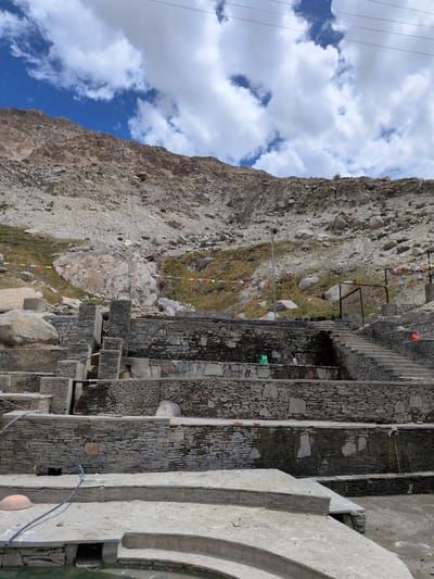 The image shows a view of the Panamik hot springs in India. The hot springs are built into the side of a mountain, with multiple tiered pools constructed from stone. The pools are filled with water and appear to be a place of relaxation or bathing. The surrounding landscape is rocky and mountainous, with sparse vegetation. The sky is partly cloudy with a bright blue sky visible in some areas. There are prayer flags strung across the hillside. The overall scene suggests a natural and spiritual destination.