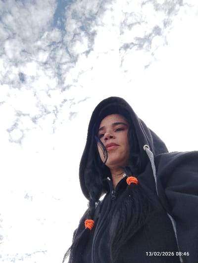 An outdoor, low-angle image depicts an adult female positioned in Puembo, Ecuador. She is shown from the upper torso upwards, wearing a dark, possibly black, hooded jacket with the hood pulled over her head. Her dark hair is styled into two pigtails or braids, each secured with a distinct orange hair tie. A single strand of hair rests across her forehead and left eye. Her lips are colored with a reddish hue. Her gaze is directed upwards and towards the right side of the frame. The background is composed almost entirely of a bright sky with numerous white and grey cumulus clouds interspersed with areas of light blue. A digital timestamp, "13/02/2026 08:46", is present in the lower right corner of the image.