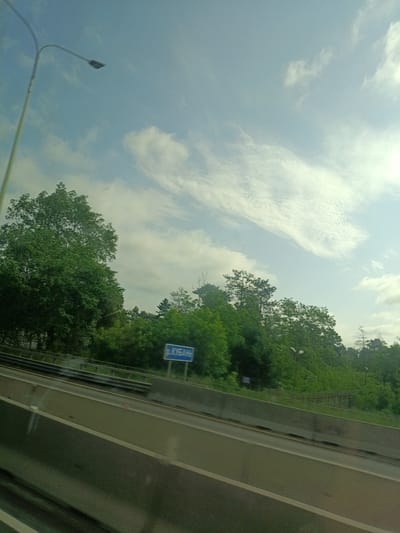 The image shows a view from a moving vehicle on a highway near Tlustenkhabl, Russia. A street light is visible on the left. The highway has a concrete barrier. Behind the barrier is a wooded area with green trees and a road sign that reads "р. КУБАНЬ". The sky is blue with white clouds.