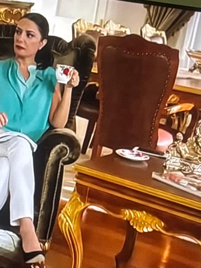 The media file shows a woman reclining on a dark couch in what appears to be a richly decorated room. She is wearing a light blue top and white pants. In her right hand, she holds a white mug with a red heart design. Her expression is relaxed, and she is looking towards the viewer. The setting is opulent, with ornate gold trim visible on a large piece of furniture in the background. A dark leather chair is also partially visible. The lighting suggests it is daytime, and the overall atmosphere is one of leisure and comfort. There is no visible text. The location context is Zarnevo, Bulgaria.