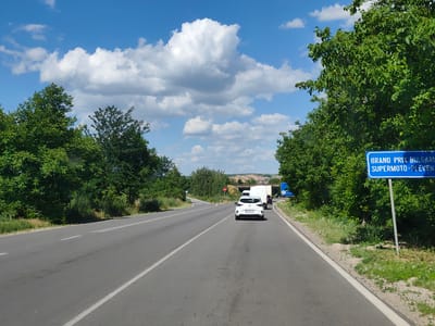 The image shows a two-lane asphalt road in a rural area. The road is bordered by lush green trees and vegetation on both sides. The sky is blue with scattered white clouds. On the right side of the road, there is a blue sign with white lettering that reads "GRAND PRIX BULGARIA SUPERMOTO-PLEVEN". A white car is driving in front of a white van on the road. The overall scene suggests a sunny day in a scenic location. The location is identified as the city of Yasen, Bulgaria.