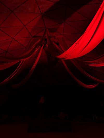 The image depicts the interior of a dome-like structure, possibly a venue or performance space in Žemaitkiemis, Lithuania. The ceiling is a geometric pattern of triangular panels, illuminated with a deep red light. Draped across the ceiling are flowing red fabrics, creating a sense of drama and visual texture. The lower half of the image is plunged into darkness, with only faint outlines of people visible. They appear to be seated or gathered on the floor, their forms silhouetted against the dim red light. The overall atmosphere is mysterious and theatrical, with the striking red lighting dominating the scene.