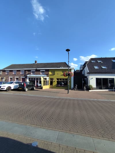 A sunny day in Schaijk, Netherlands, is depicted in this image of a street scene. The sky is a vibrant blue with scattered white clouds, suggesting pleasant weather. The foreground is a brick-paved street, with a gray concrete curb and sidewalk.

Along the street are several buildings. On the left is a row of brick buildings with multiple windows and dark roofs. Parked in front of these buildings are two cars: a white Audi SUV and a black small hatchback.

Next to the brick buildings is a bright yellow building with large storefront windows. A sign above the entrance reads "Sisters." Another building, painted white, is to the right of the yellow one, featuring a multi-level roof with several skylights and large glass doors.

A tall lamppost stands in front of the yellow and white buildings, with a hanging basket of pink and red flowers near its top. Two bicycles are parked near the white building. The overall scene suggests a quiet commercial area during daylight hours, possibly late morning or early afternoon given the bright sunlight and clear sky. No people are visibly present in the image.