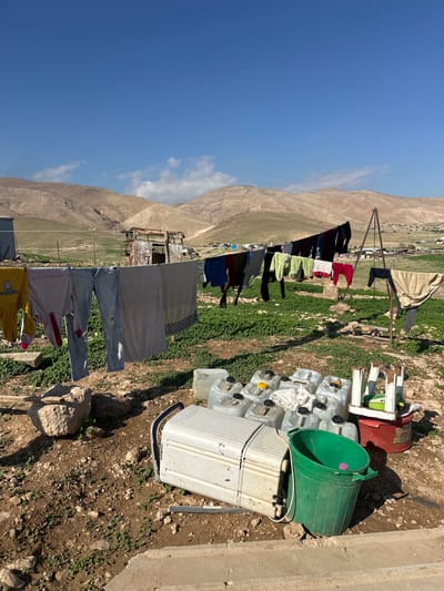 The image depicts an outdoor scene in the Arvot Hayarden Regional Council, Palestinian Territory, under a clear blue sky with minimal cloud cover. In the foreground, dry, rocky ground with sparse green vegetation is visible. A concrete edge runs along the bottom.
In the midground, a clothesline stretches across the frame, supporting various items of laundry, including trousers, shirts, and a patterned cloth, drying in the sunlight. A second clothesline, supported on the right by a tripod-like metal structure, also holds drying garments. To the right of the lower clothesline, a cluster of clear plastic jerrycans with yellow and black caps is present, accompanied by a green plastic bucket and a white, overturned object resembling a cooler or small appliance. A red plastic tub topped with a small, light-colored stand is positioned further right.
Towards the left midground, a segment of a light-colored rectangular structure is visible, along with a dilapidated, rusty corrugated metal shack featuring an open doorway. The background is dominated by a range of arid, light-brown mountains with sparse green patches on their lower slopes. Distant structures, possibly tents or small buildings, are scattered in the valley between the foreground and the mountain range. Shadows are cast to the right of objects, indicating a strong light source from the left. No individuals are present in the scene.