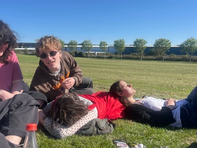 The image depicts a group of young people relaxing on a sunny day in a grassy field in Dundee, United Kingdom. The sky is a clear, vibrant blue, indicating fair weather.
In the foreground and mid-ground, several individuals are visible. On the far left, a person with dark, curly hair and a pink striped shirt is partially seen. Central to the frame, a young person with short, curly light brown hair, wearing sunglasses and a brown hoodie, smiles towards the viewer, holding a small item in their hands. To their right, another individual with dark curly hair is lying face down on the green grass, dressed in a white top with black polka dots. A grey utility pouch rests beside them. Further to the right, a young woman lies on her back on a bright red fabric, with her eyes closed, seemingly enjoying the sun. She wears a white top under a black long-sleeved garment, with blue jeans. Part of another person's legs is visible on the far right.
The setting is a large, well-maintained grassy area. In the background, a neat row of small, deciduous trees with green foliage is evenly spaced along a fence line. Behind these trees, a series of modern, large industrial or commercial buildings, characterized by their grey and blue facades, stretch across the horizon, suggesting an area near an industrial park or commercial estate.
The activity is one of relaxation and sunbathing. The strong sunlight casts clear shadows on the grass, indicating it is midday or early afternoon. A clear plastic bottle with a red cap is visible in the bottom left corner. The overall mood is relaxed and peaceful, with individuals enjoying the warmth and sunshine. No discernible text is visible on clothing or objects.