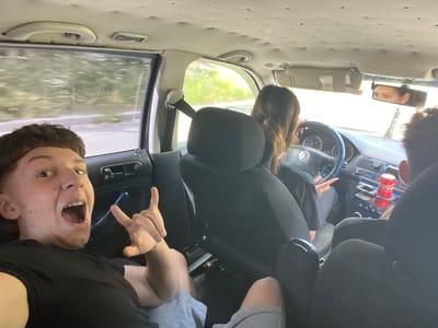 Four individuals are captured inside a moving vehicle, likely a Volkswagen, traveling through Vlahovo, Bulgaria. In the foreground, a young person with dark hair and braces is enthusiastically posing for the camera, their mouth wide open and one hand giving a peace sign. To their right, another person with dark hair is driving, their hands on the steering wheel. In the background, partially obscured by seats, two other individuals are visible, one in the passenger seat and another in the rear. A red Coca-Cola can rests in the center console, illuminated by the natural light filtering through the windows. The car's interior appears worn, with visible imperfections on the ceiling. The exterior scenery suggests a rural or suburban setting with lush greenery.